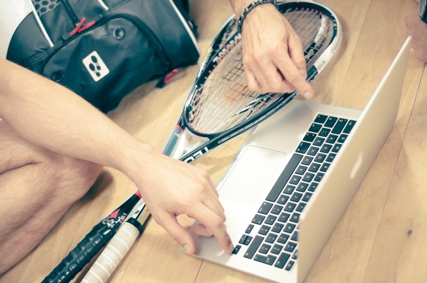 Two squash players with their equipment gathered around a laptop computer.