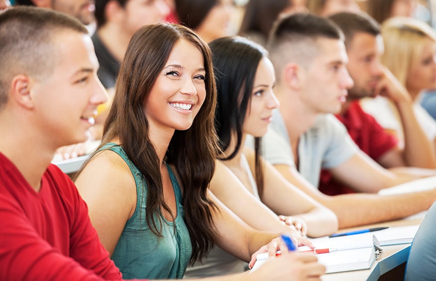 A female student smiling while in a lecture hall with many other students.