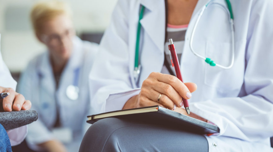 A medical professional sitting with a notebook on her lap and a stethoscope around her neck.