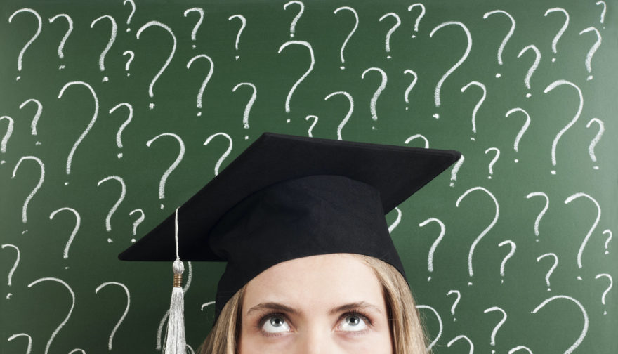 A graduating senior in her cap standing in front of a chalk board covered in question marks.