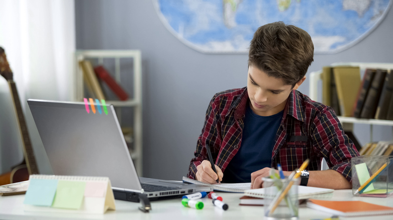 A young student taking notes in a notebook from information gained on his laptop.