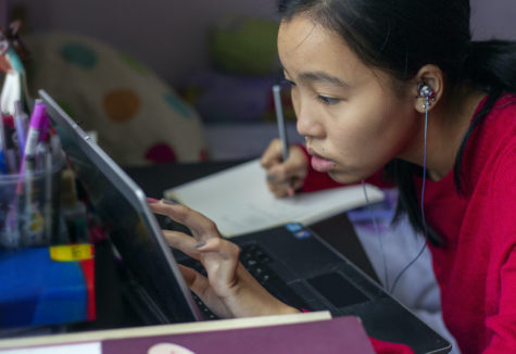 A female student of Asian descent studying hard at her laptop while taking notes in a notebook.