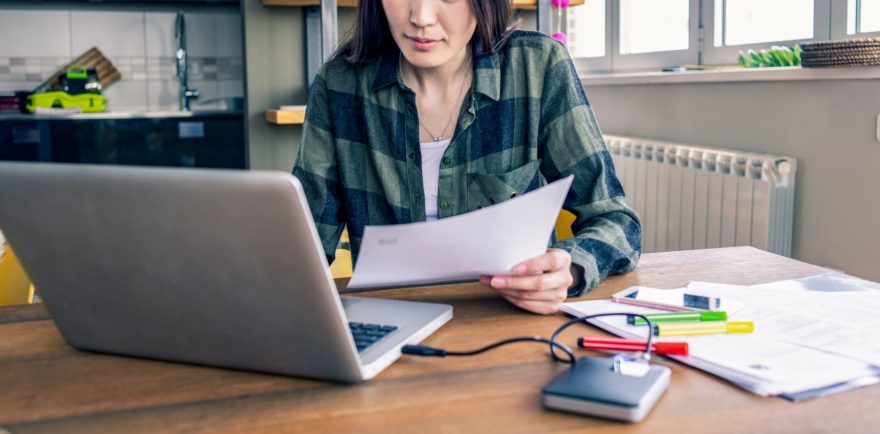 A woman working in front of a laptop, surrounded by papers, colored pens, and an external hard-drive.