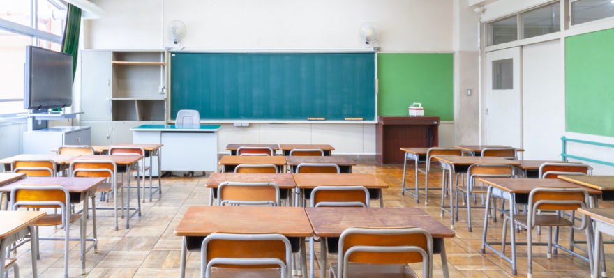 The inside of an empty classroom with a chalkboard and wooden-topped desks.