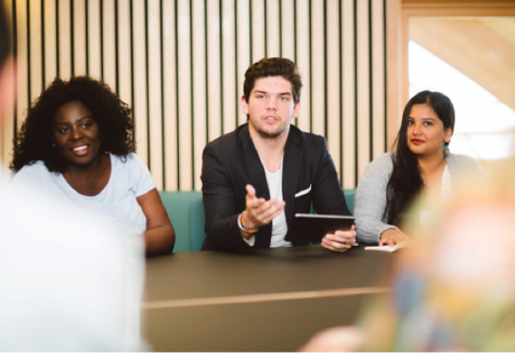 Three students, two female one male, presenting something to a group from across a conference table.