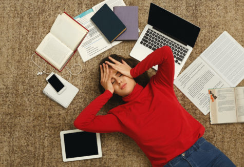 A student laying down, surrounded by textbooks, study electronics, and notebooks as she holds her head in stressful frustration.