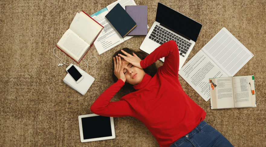 A student laying down, surrounded by textbooks, study electronics, and notebooks as she holds her head in stressful frustration.