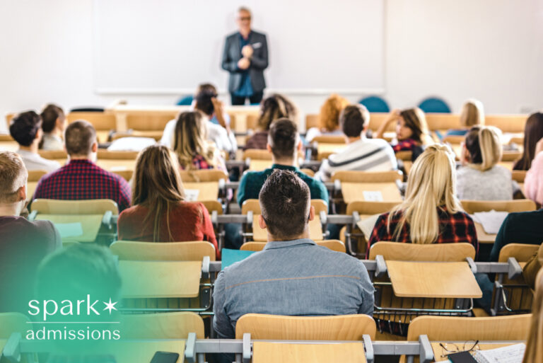 College students attending a university lecture in a large classroom while a professor teaches at the front of the room.