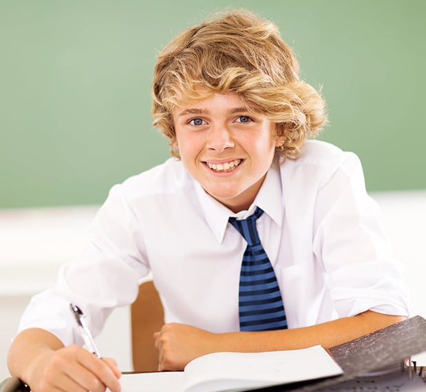 Smiling young blonde teenage boy in private school uniform and tie sitting at desk with chalkboard behind him