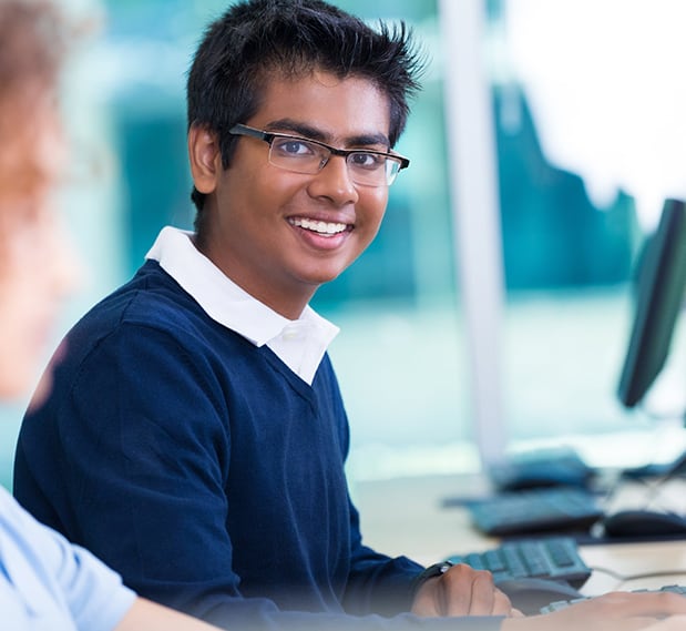 Smiling teen boy of Indian descent wearing school uniform seated in front of computer in computer lab