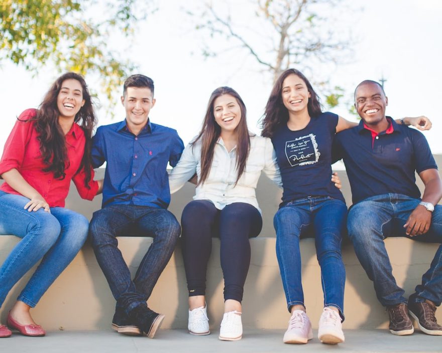 A set of five friends or roommates sitting together on a low wall.