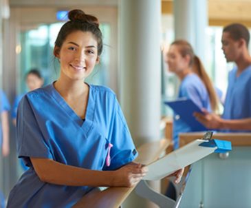 Smiling female med student in scrubs standing at nurse’s station with other people in scrubs behind her