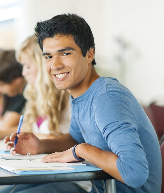 Smiling teen boy of Indian descent writing in notebook at desk in classroom