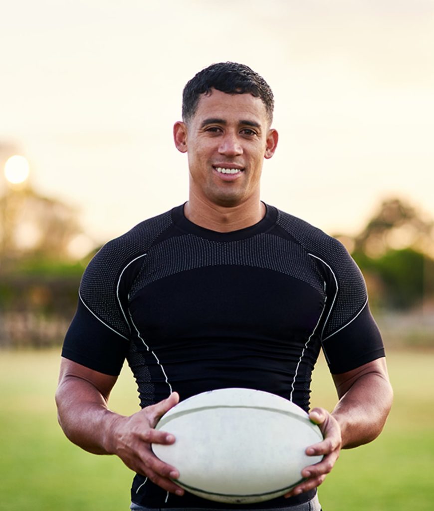 A college male athlete holding a rugby ball on a field and smiling at the camera