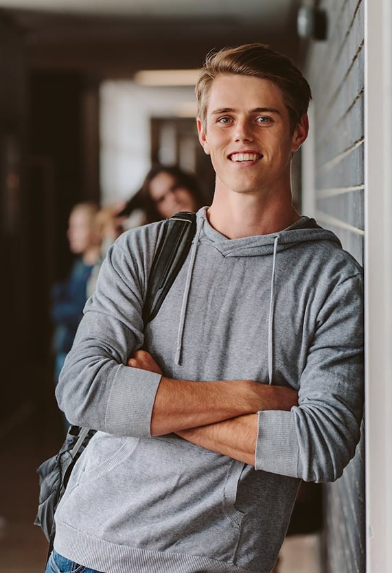 Tall high school boy with blonde hair leaning against hallway wall with arms crossed and other students in background