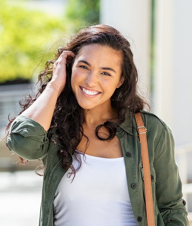 Smiling teen girl with dark curly hair standing outside school tucking hair behind ear