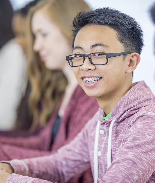 Smiling young high school boy of Asian descent wearing glasses and braces sitting at desk in classroom with classmates behind him
