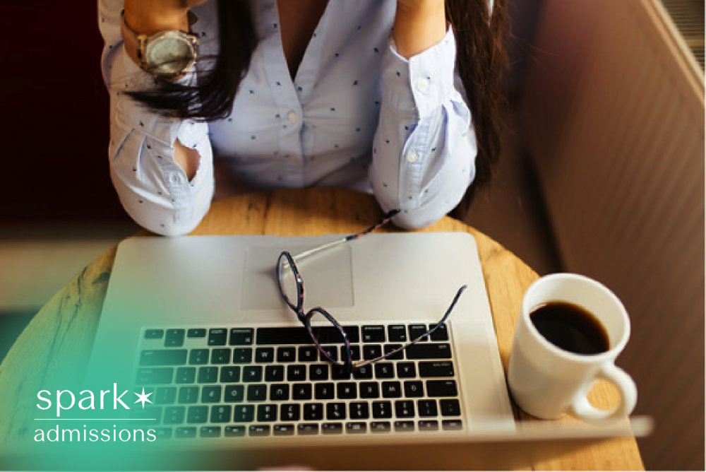 Student sitting at a laptop with coffee and eyeglasses, looking stressed while reviewing coursework or preparing for an online exam.