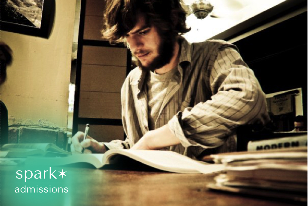 College student writing notes in an open textbook while studying at a desk surrounded by books in a quiet study space.