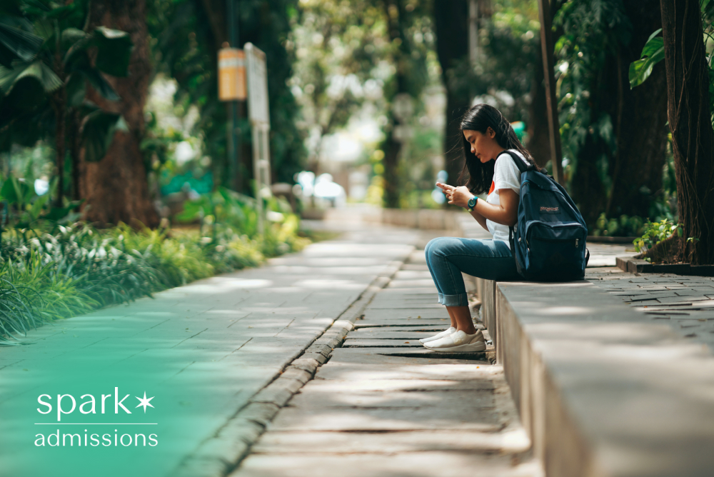Student with backpack using a smartphone while sitting on a campus walkway surrounded by trees.