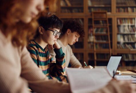 Students studying at a library table with books and a laptop, focused on schoolwork.