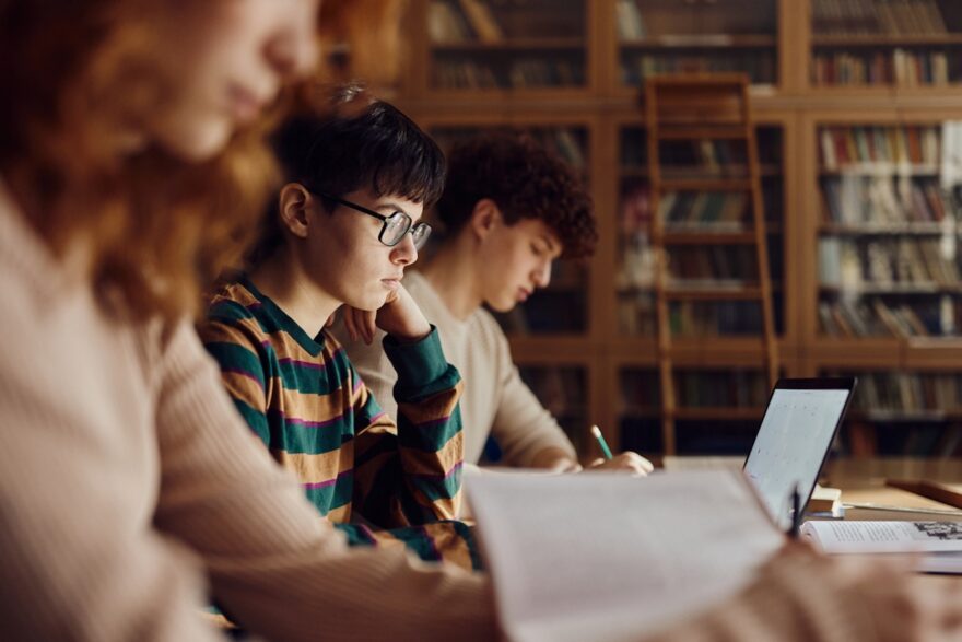 Students studying at a library table with books and a laptop, focused on schoolwork.