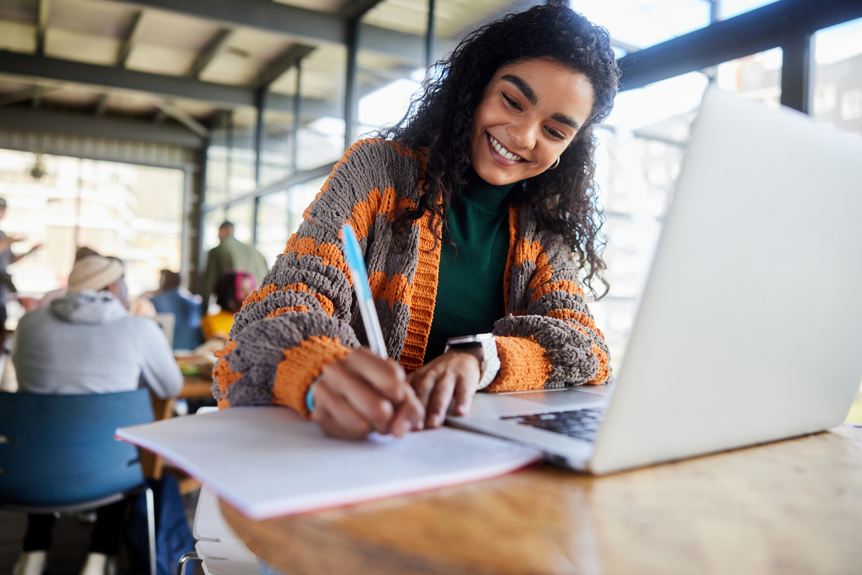 College student writing notes while studying with a laptop in a campus workspace.