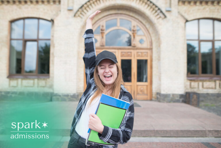 Happy college student celebrating outside a university building while holding notebooks, raising one arm in excitement after academic success or college acceptance.