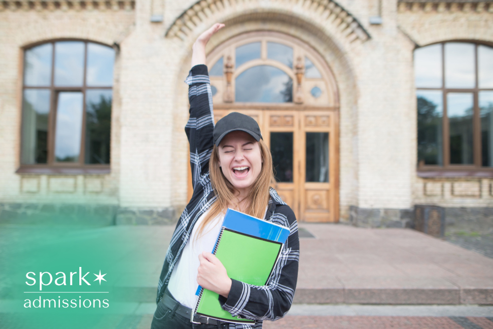 Happy college student celebrating outside a university building while holding notebooks, raising one arm in excitement after academic success or college acceptance.