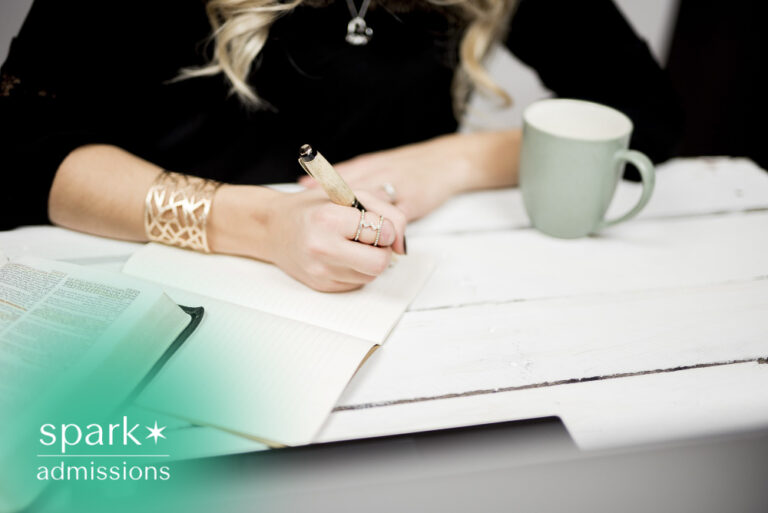 Student writing notes in a notebook during a study session at a desk with an open book and coffee mug, representing college admissions preparation and academic success.