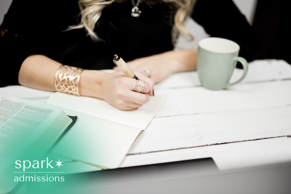 Student writing notes in a notebook during a study session at a desk with an open book and coffee mug, representing college admissions preparation and academic success.