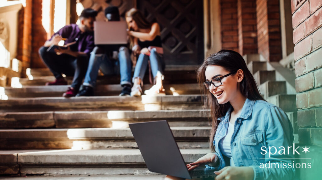 A student sitting on outdoor stairs, smiling while using a laptop.