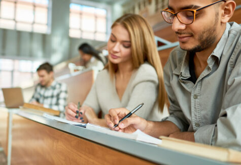 Multi-ethnic group of students sitting at desk in lecture hall of modern college and writing, focus on young Middle-Eastern man wearing glasses, copy space