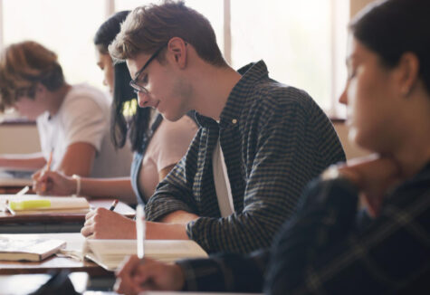 Focused student sitting at a desk, writing answers on an exam sheet in a classroom setting.