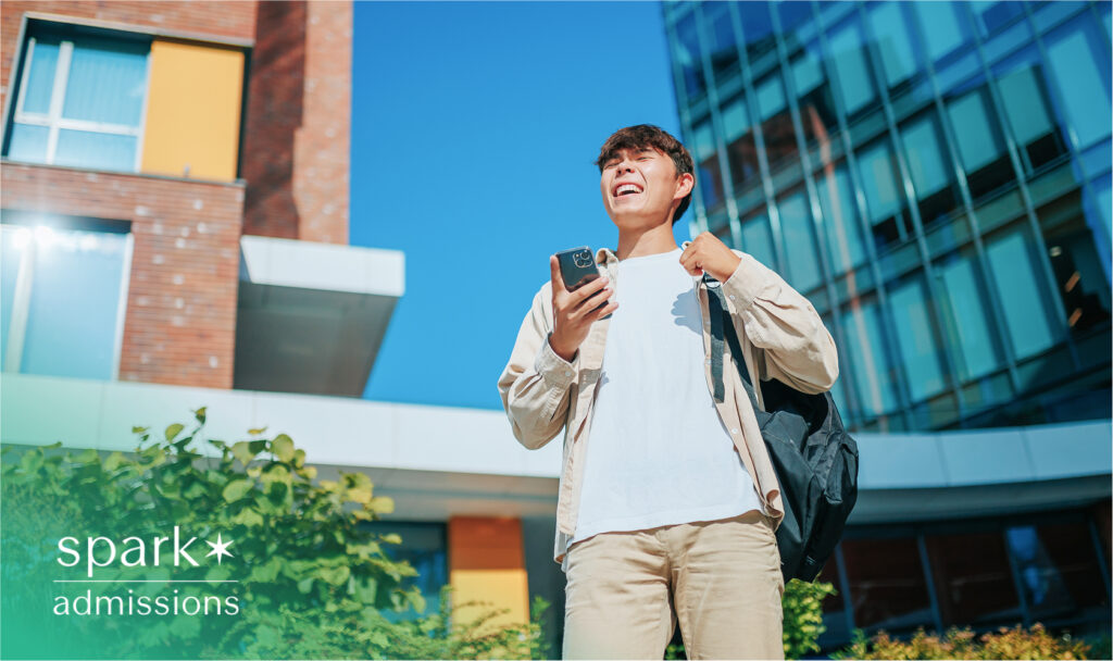 Smiling student holding phone outside school building, looking ahead confidently