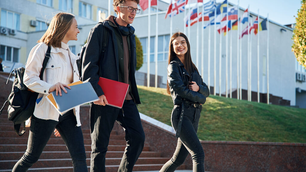 Three students walking down steps outside a university with international flags in the background
