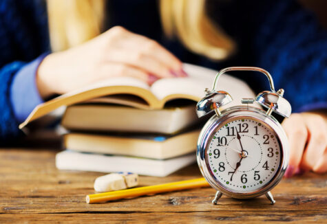 Blurred student studying in the background with a focused alarm clock in front, symbolizing waiting for college acceptance