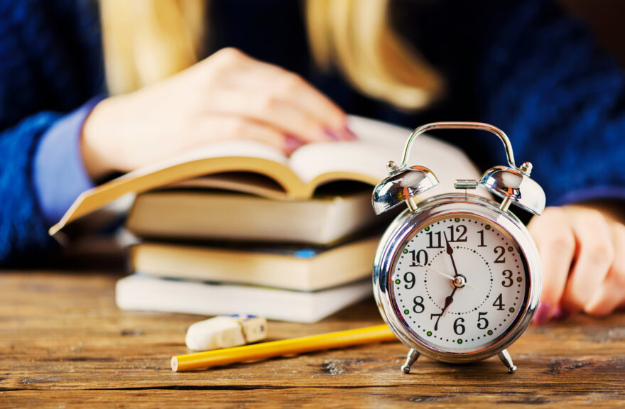 Blurred student studying in the background with a focused alarm clock in front, symbolizing waiting for college acceptance