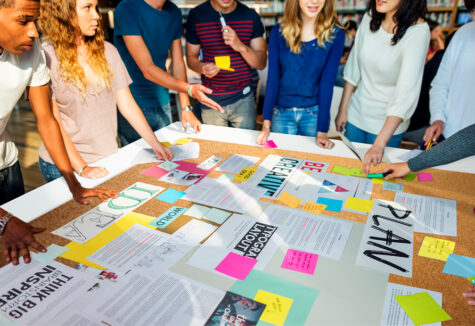 Group of students gathered around a large table filled with notes, colorful papers, and planning materials, actively collaborating and organizing ideas for a project