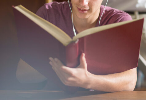 Student standing in a library, holding a large red book while studying, surrounded by shelves filled with books, focused and engaged in learning.