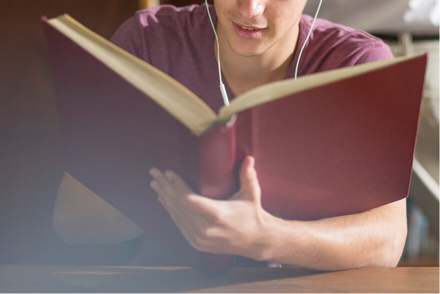 Student standing in a library, holding a large red book while studying, surrounded by shelves filled with books, focused and engaged in learning.
