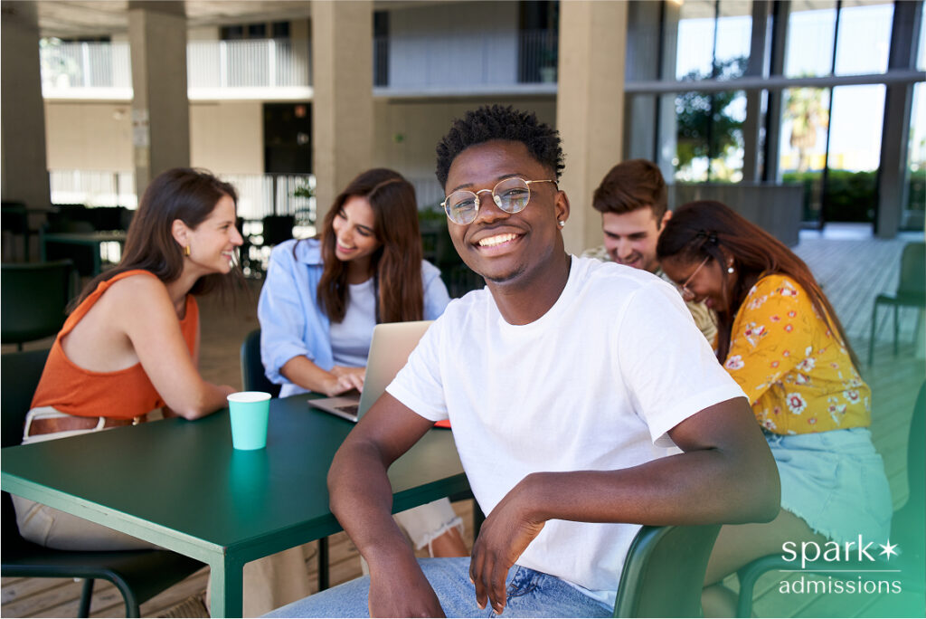 Confident student smiling at the camera while sitting with a diverse group of peers engaged in conversation and study, representing academic support and success with Spark Admissions.