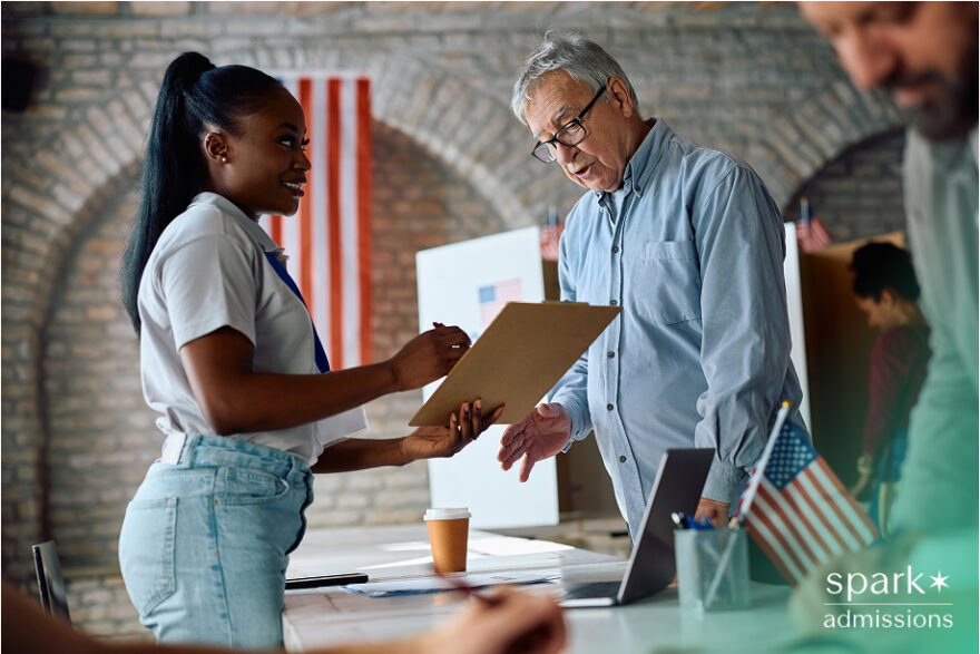 A student hands a clipboard to an older man at a volunteer or civic event table