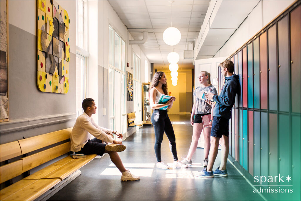Four students talk near lockers in a school hallway while one student sits on a bench