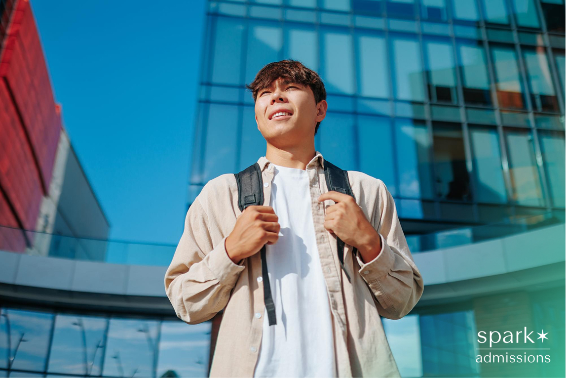 A male student wearing a backpack gazes ahead confidently while standing in front of a modern glass academic building