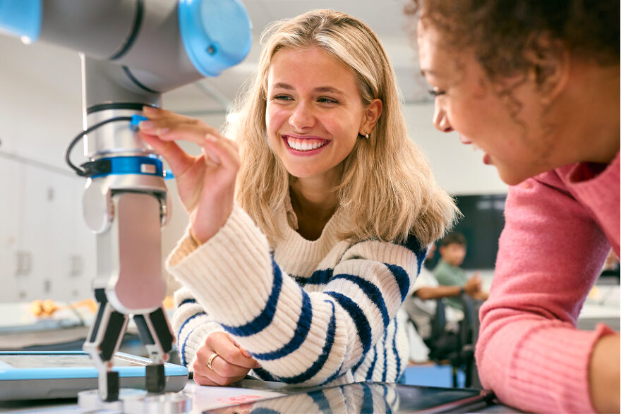 Two Female College Or University Engineering Students Using Digital Tablet To Control Robot Arm