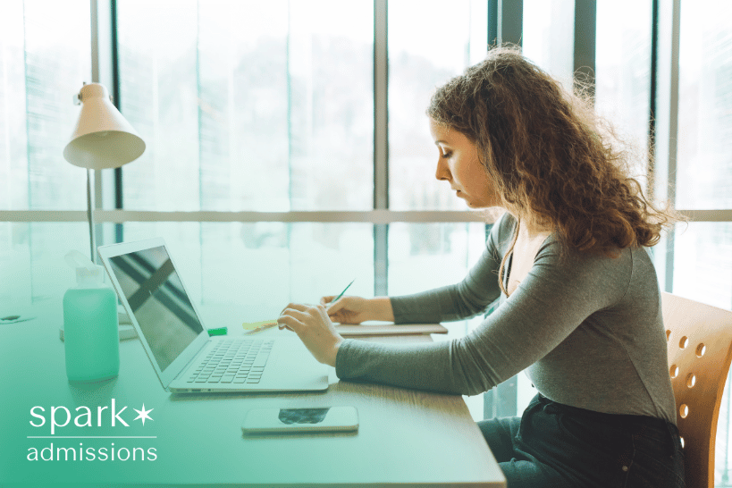 High school student writing an essay for college admission on a desk with books and laptop