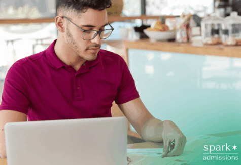 Man wearing glasses writing a transfer essay on laptop while reviewing notes on paper