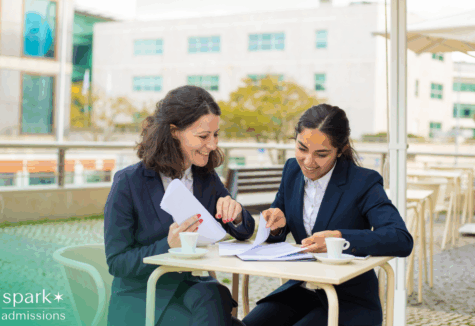 Two female business college students smiling while reviewing papers together at a café table