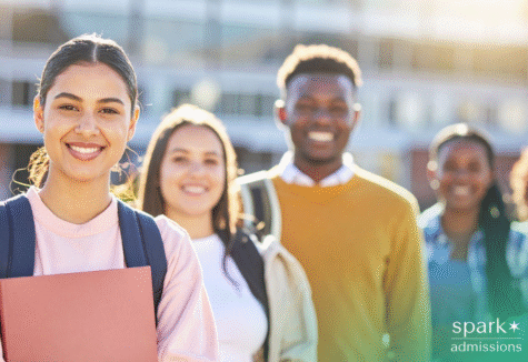 Diverse group of smiling college students standing outdoors on campus, with one student holding a folder in the foreground.